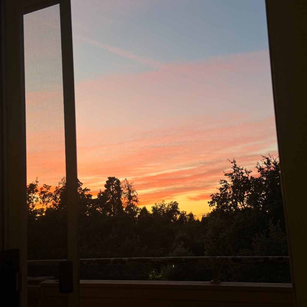 Sunset sky fading from peach to blue, framed by a window and silhouetted trees