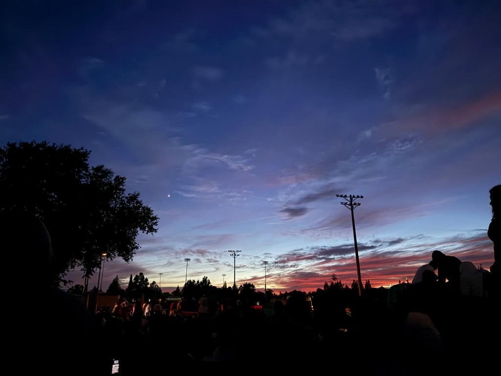 Large crowd silhouetted against a vibrant blue and orange sunset sky at an outdoor field with tall stadium lights